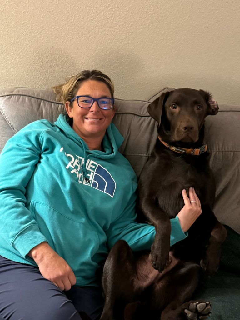 A woman is sitting on the couch with her chocolate lab